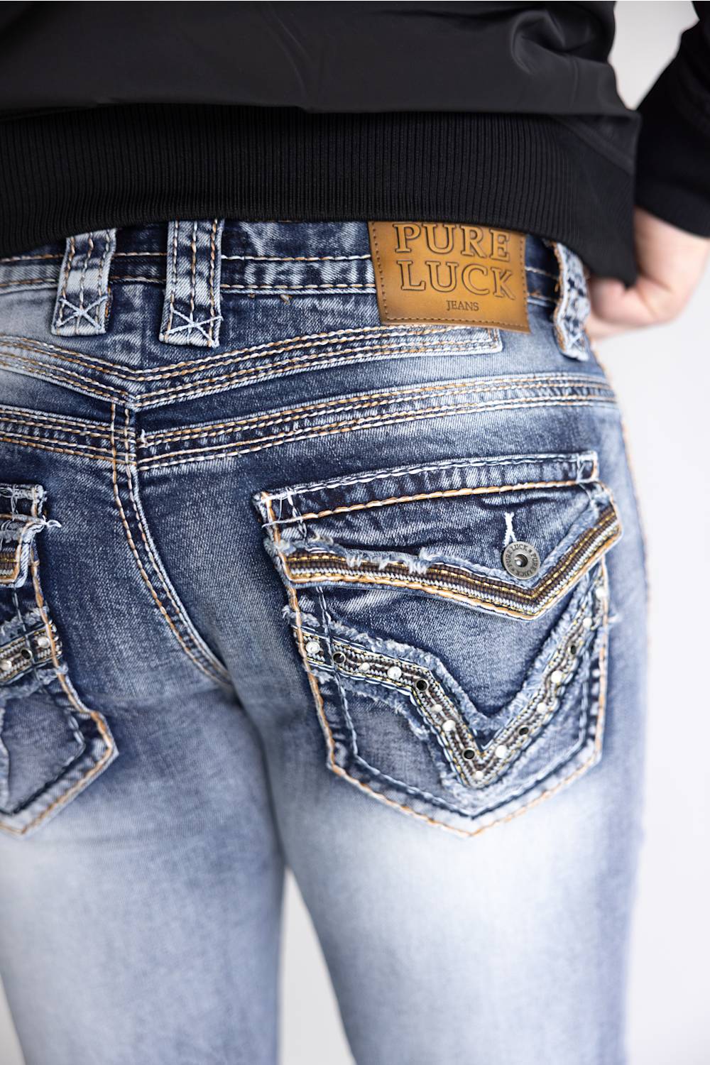 Close-up back view of a person wearing blue denim jeans with a tan leather Pure Luck brand patch at the waistband, paired with a black top against a light gray background.