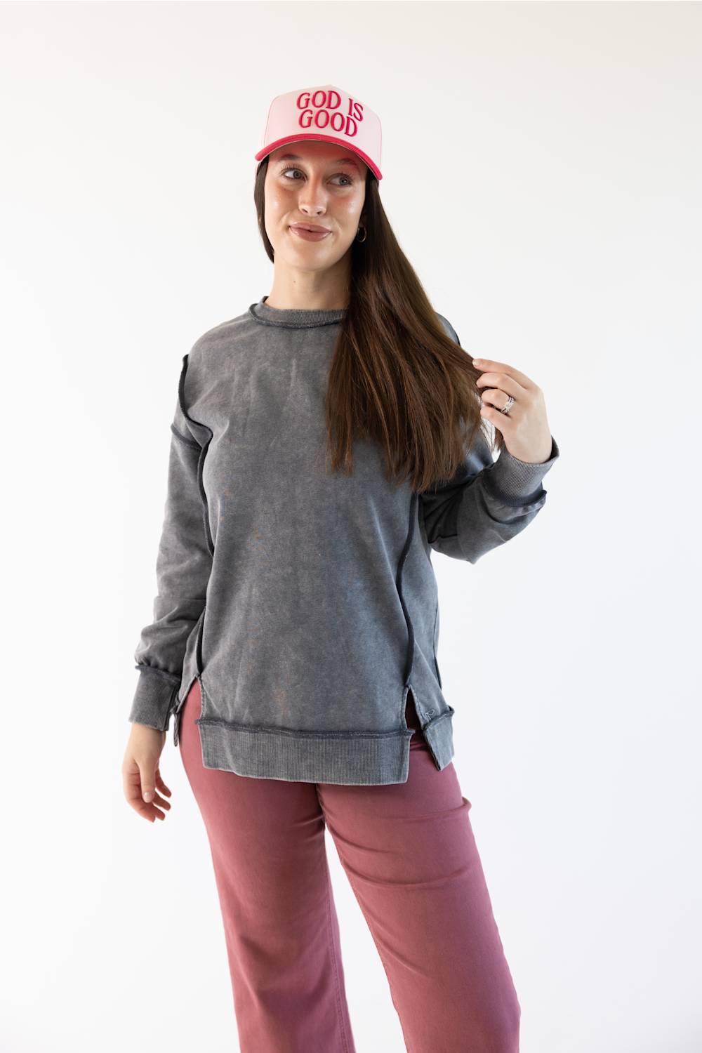 A woman modeling a gray long-sleeve crewneck sweatshirt with side slits, paired with dusty rose pants and a pink cap that reads 'GOD IS GOOD', against a plain white background.