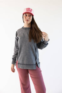 A woman modeling a gray long-sleeve crewneck sweatshirt with side slits, paired with dusty rose pants and a pink cap that reads 'GOD IS GOOD', against a plain white background.