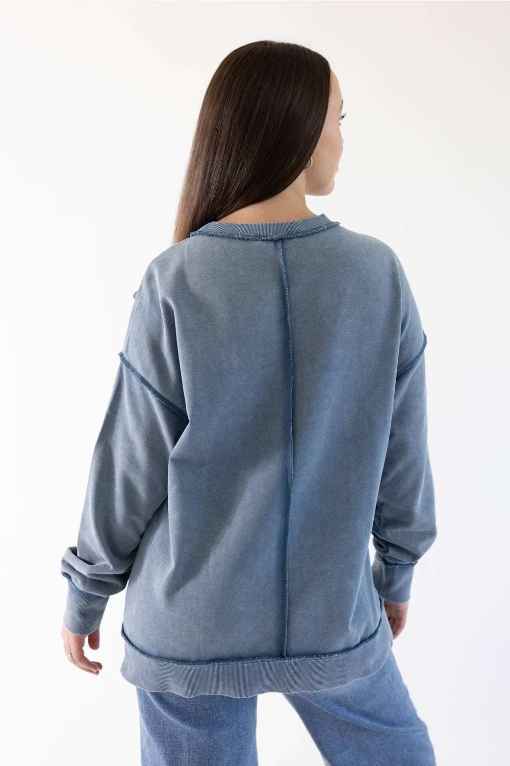 A woman viewed from the back wearing a washed slate blue sweatshirt and matching blue jeans against a plain white studio background.