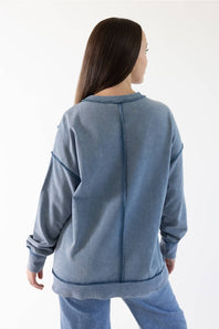 A woman viewed from the back wearing a washed slate blue sweatshirt and matching blue jeans against a plain white studio background.