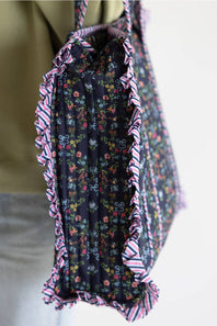 A close-up of a fabric tote bag being carried on a person's shoulder. The bag has a dark floral print with pink, red, and blue flowers, and is finished with pink, white, and blue striped ruffled trim.