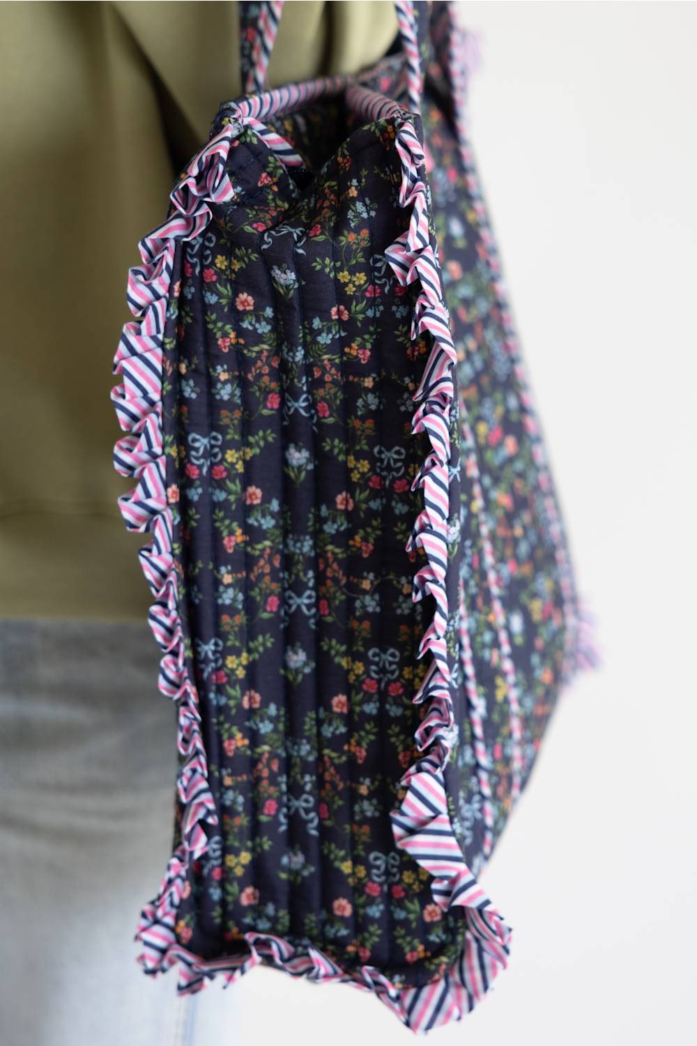 A close-up of a fabric tote bag being carried on a person's shoulder. The bag has a dark floral print with pink, red, and blue flowers, and is finished with pink, white, and blue striped ruffled trim.