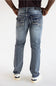 Rear view of a person wearing light-wash blue denim jeans with decorative stitching and silver-toned hardware on the back pockets, paired with a dark top and white sneakers.