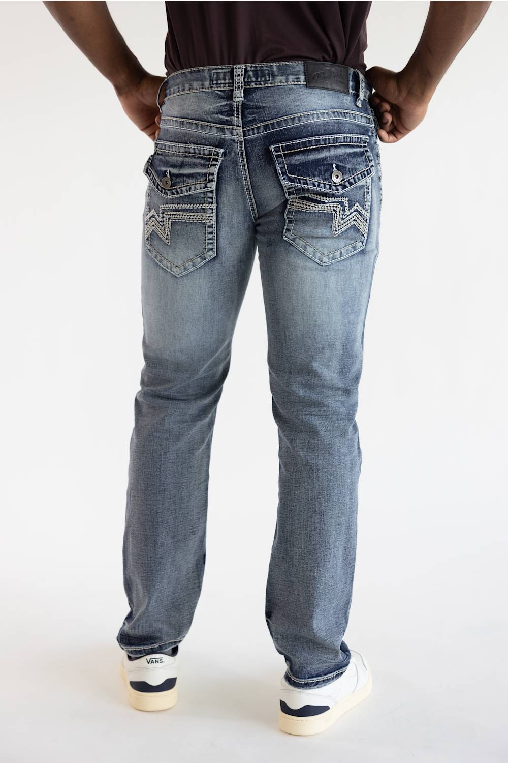 Rear view of a person wearing light-wash blue denim jeans with decorative stitching and silver-toned hardware on the back pockets, paired with a dark top and white sneakers.
