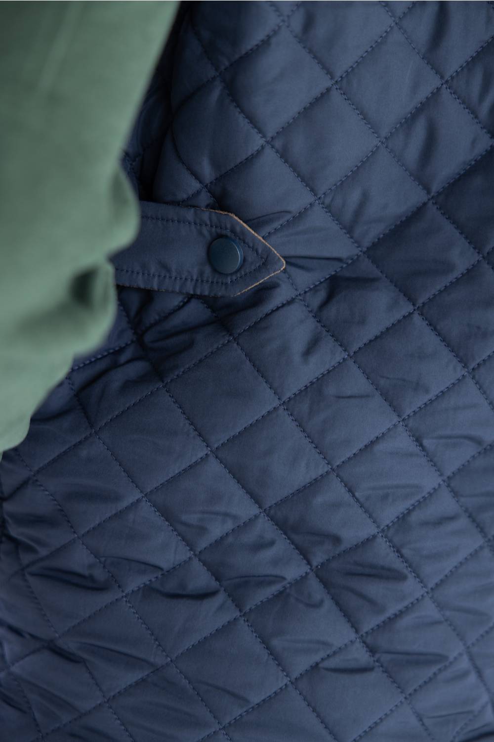 Close-up of a navy blue quilted jacket with a diamond-stitched pattern and a flap pocket with a snap closure, alongside a muted green fabric section at the left edge.