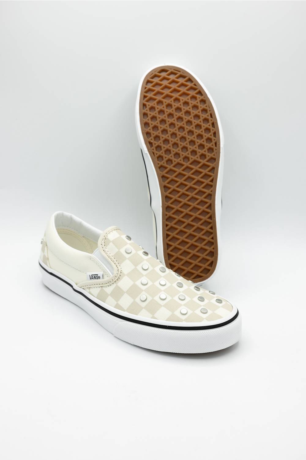 A pair of Vans slip-on sneakers in off-white with a checkerboard pattern and silver studs, with one shoe standing upright to show the brown waffle outsole.