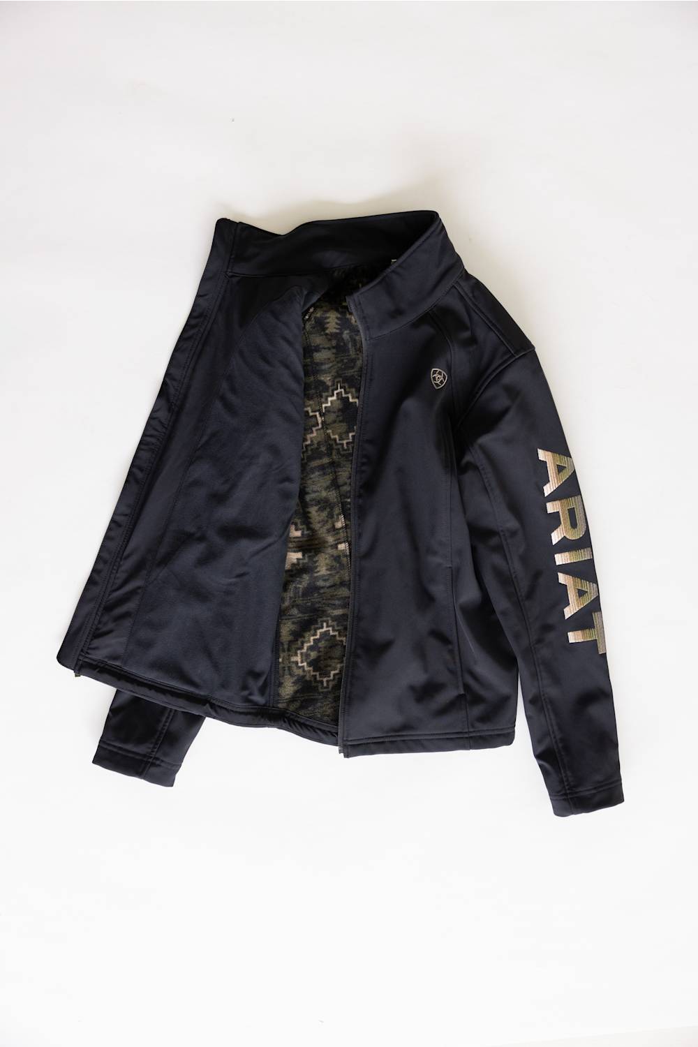 A black zip-up jacket with a high collar, laid flat on a white background. The jacket features a camouflage-patterned inner lining visible at the open front, a small circular logo on the left chest, and the brand name 'ARIAT' printed vertically in gold on the right sleeve.