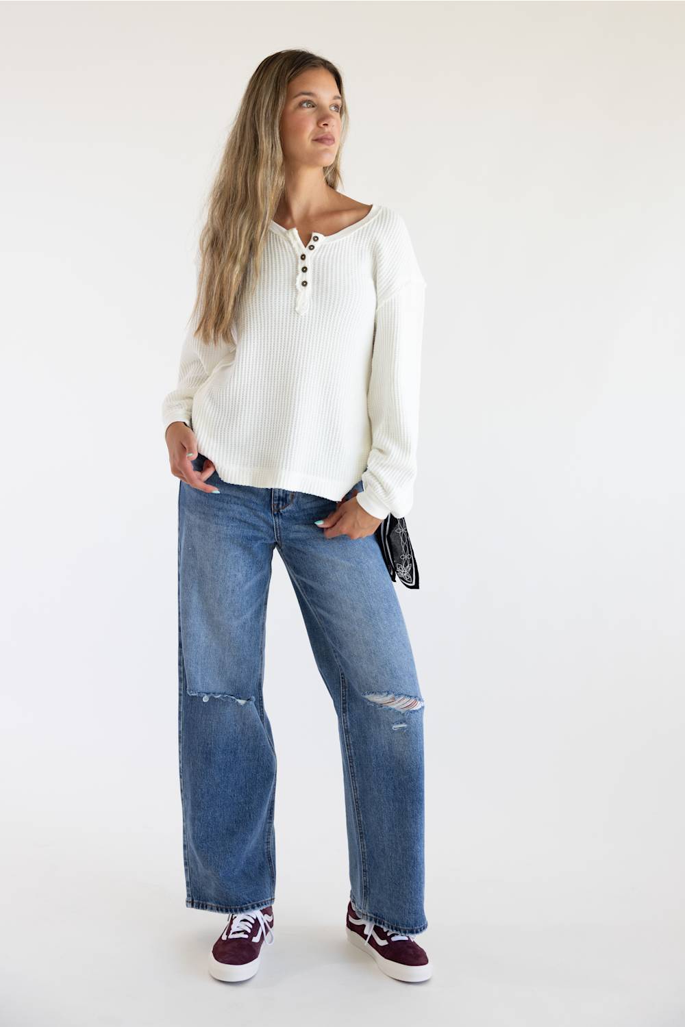 A woman modeling a white ribbed long-sleeve henley top with blue wide-leg distressed jeans and burgundy sneakers, standing against a plain white studio background.