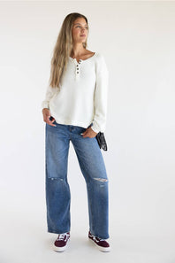 A woman modeling a white ribbed long-sleeve henley top with blue wide-leg distressed jeans and burgundy sneakers, standing against a plain white studio background.