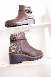 A pair of women's brown ankle boots with a textured knit pattern panel and a buckle strap, displayed on white wooden shelves.
