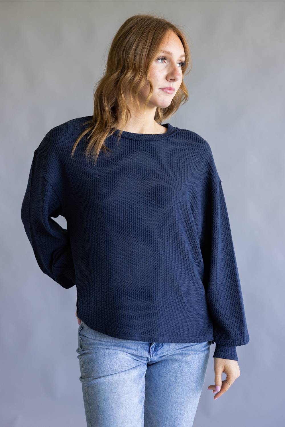 A woman wearing a dark navy blue waffle-knit long-sleeve sweater and light blue jeans, standing against a neutral gray studio background.
