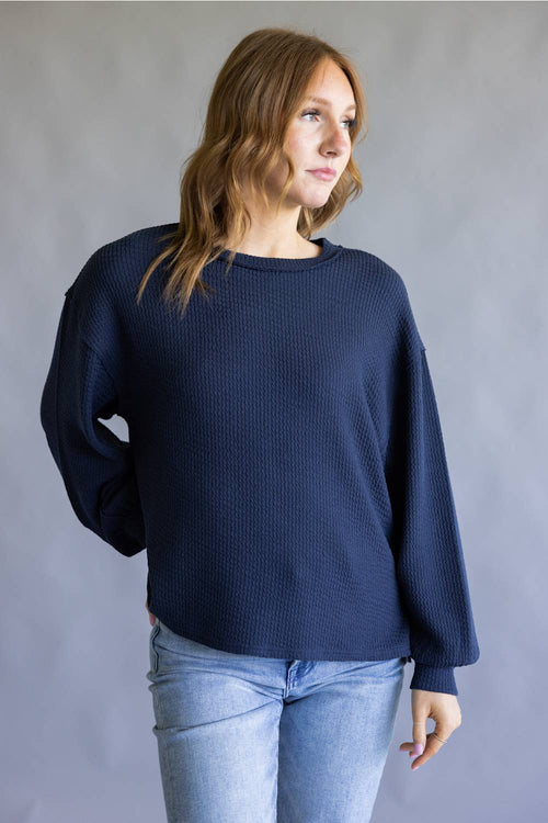 A woman wearing a dark navy blue waffle-knit long-sleeve sweater and light blue jeans, standing against a neutral gray studio background.