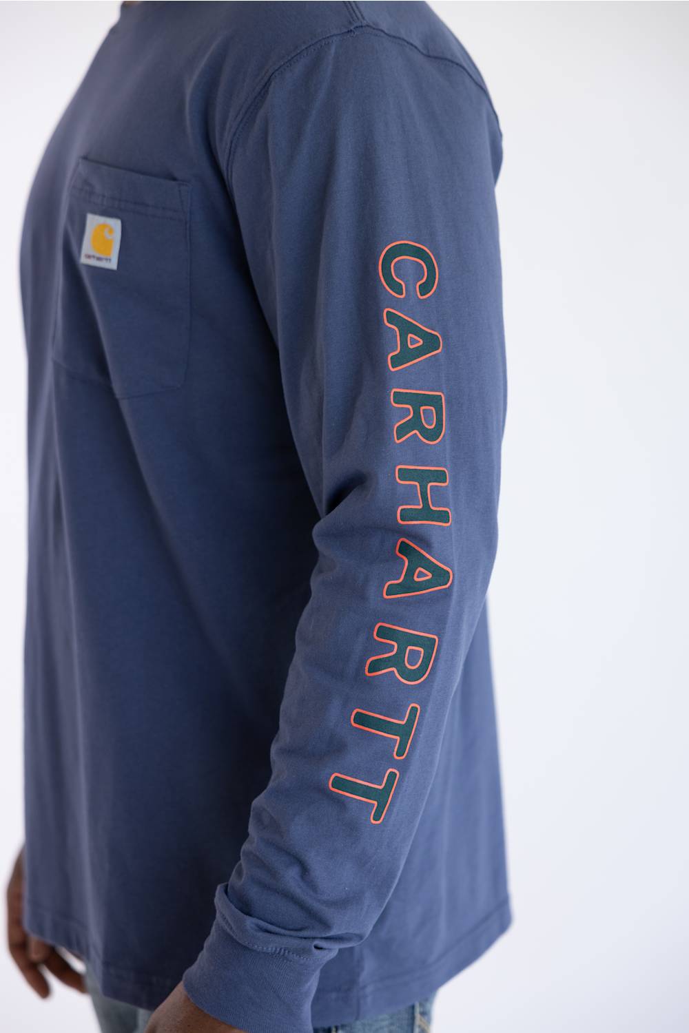 A close up from the side of a man wearing a long sleeve Carhartt t-shirt in Superstorm blue with a classic Carhartt patch on the front chest pocket located on the upper left with a Carhartt logo down the left sleeve.