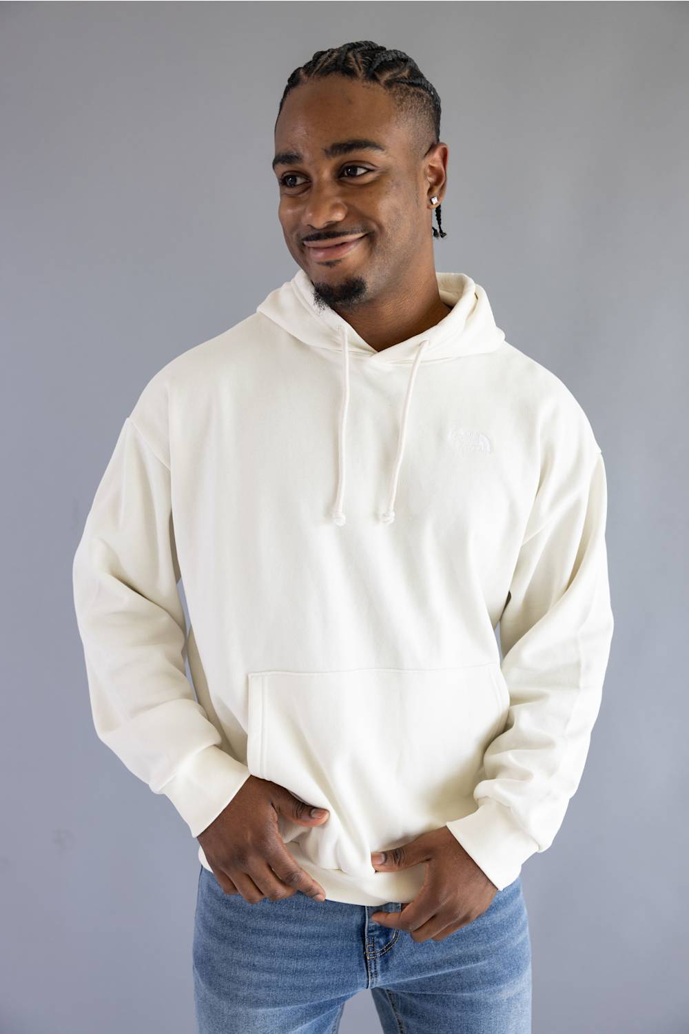 A man wearing an off-white pullover hoodie with drawstrings and a front kangaroo pocket, paired with blue jeans, standing against a solid gray studio background.