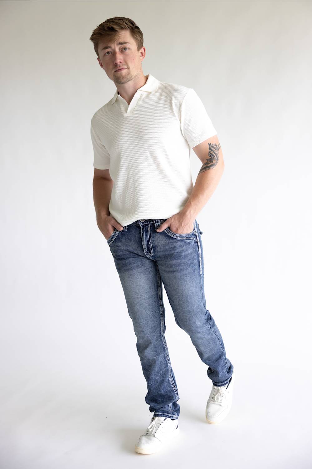 A man wearing a white short-sleeve polo shirt, blue jeans, and white sneakers, standing against a plain light gray studio background.