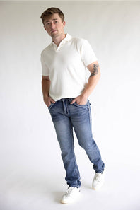 A man wearing a white short-sleeve polo shirt, blue jeans, and white sneakers, standing against a plain light gray studio background.