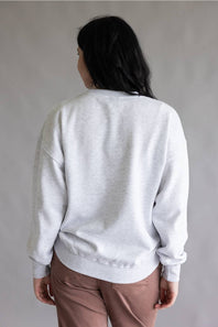 A person shown from the back wearing a light gray heather crewneck sweatshirt and dusty rose pants, standing against a neutral gray studio background.