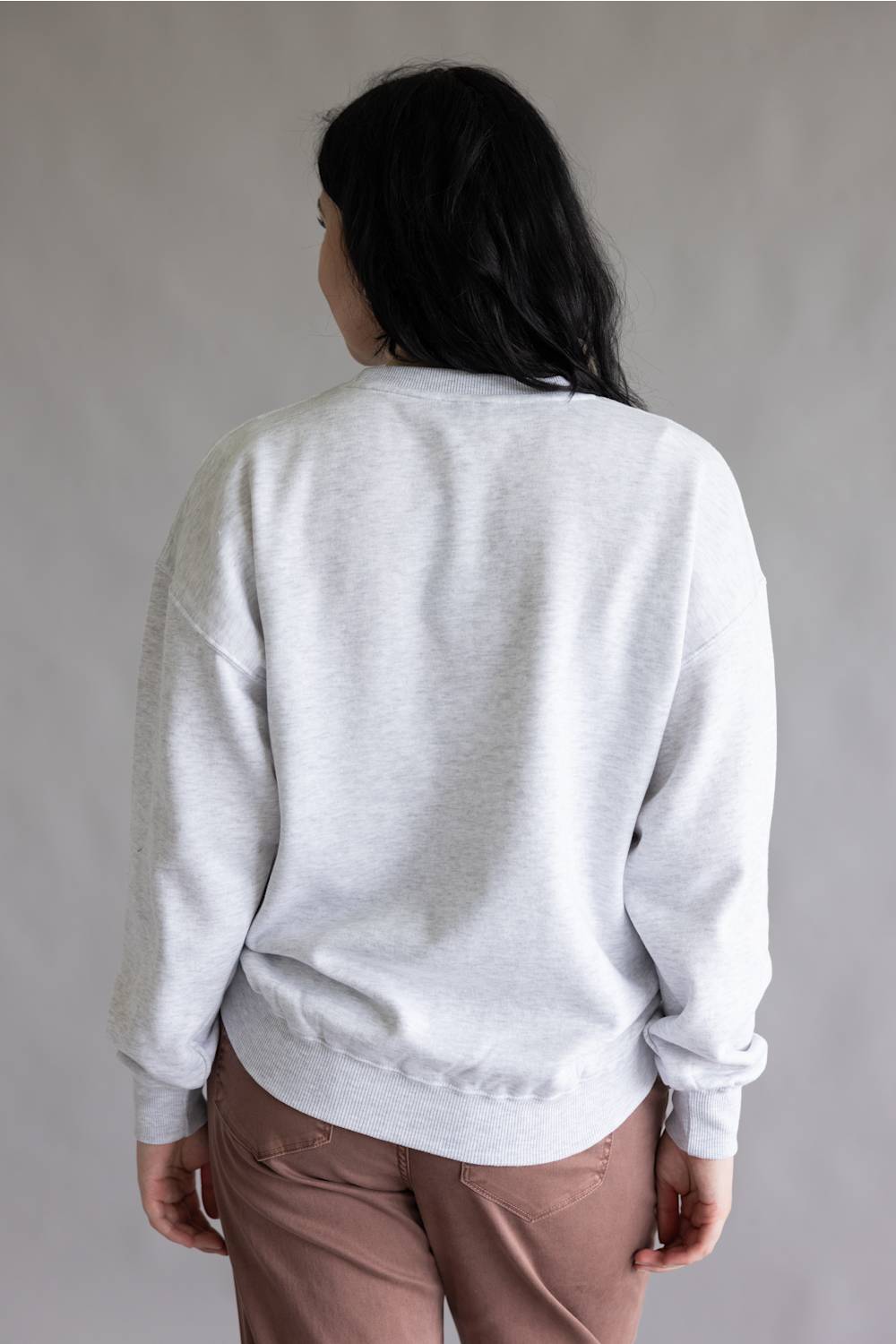A person shown from the back wearing a light gray heather crewneck sweatshirt and dusty rose pants, standing against a neutral gray studio background.
