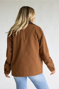 A woman shown from the back wearing a brown utility-style jacket and light blue jeans against a plain light gray studio background.