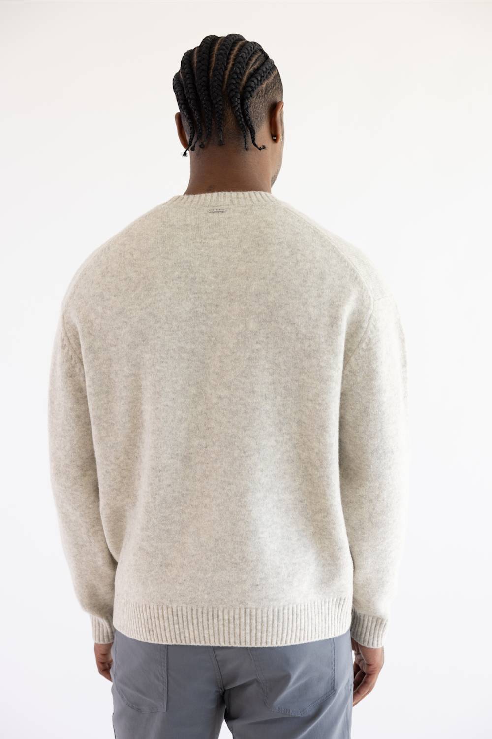 A man viewed from behind wearing a light beige knit crewneck sweater and gray trousers, standing against a plain white studio background.