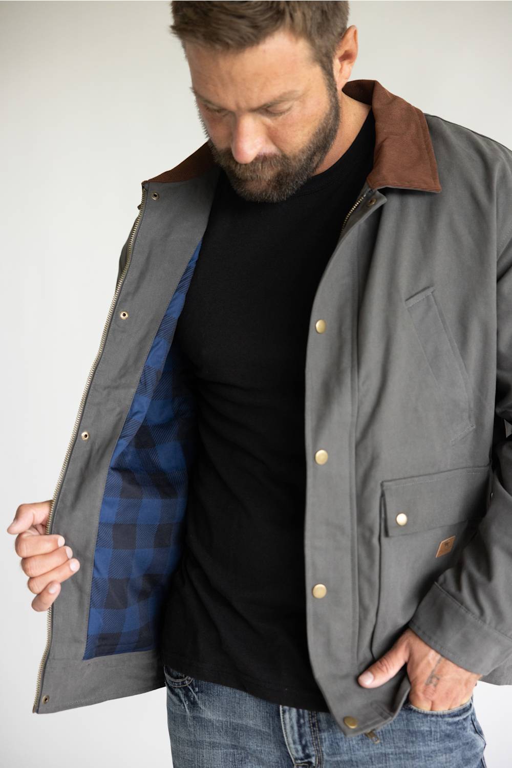 A man wearing a gray waxed cotton utility jacket with a brown corduroy collar, styled over a black t-shirt and blue jeans, standing against a light gray studio background.