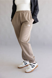 A person wearing beige jogger sweatpants with an elastic waistband and side pocket, styled with a white top, black jacket, and white sneakers with black stripes, against a plain gray studio background.