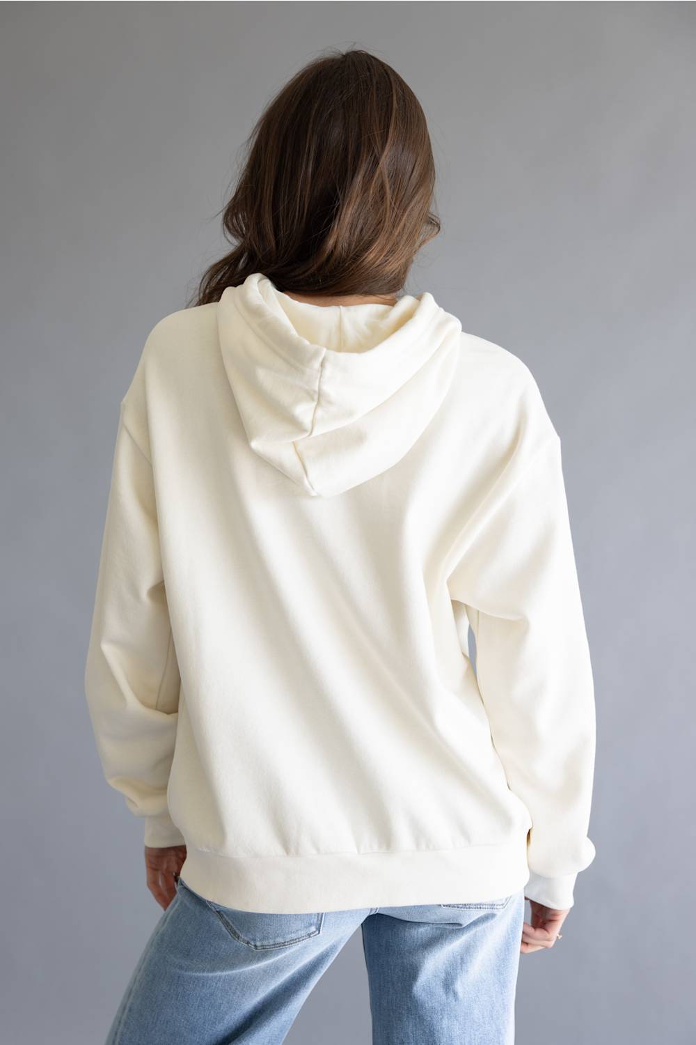 A person viewed from the back wearing an oversized cream-colored hoodie and light blue jeans, standing against a plain gray studio background.