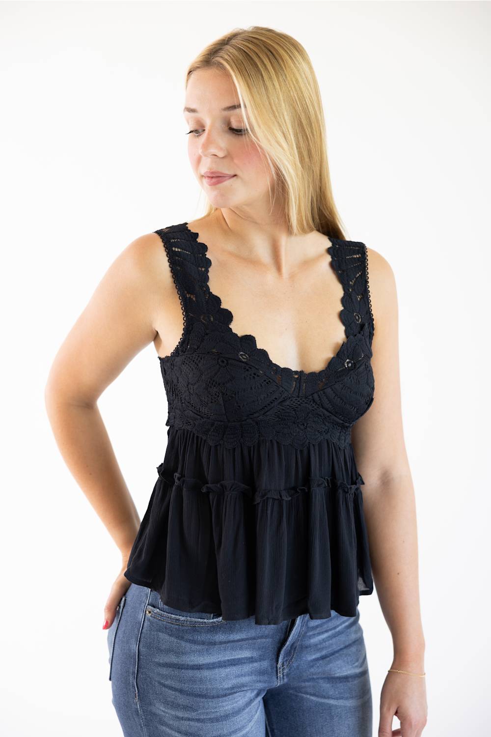 A woman wearing a black sleeveless lace-trimmed ruffled top and blue jeans, standing against a plain white studio background.