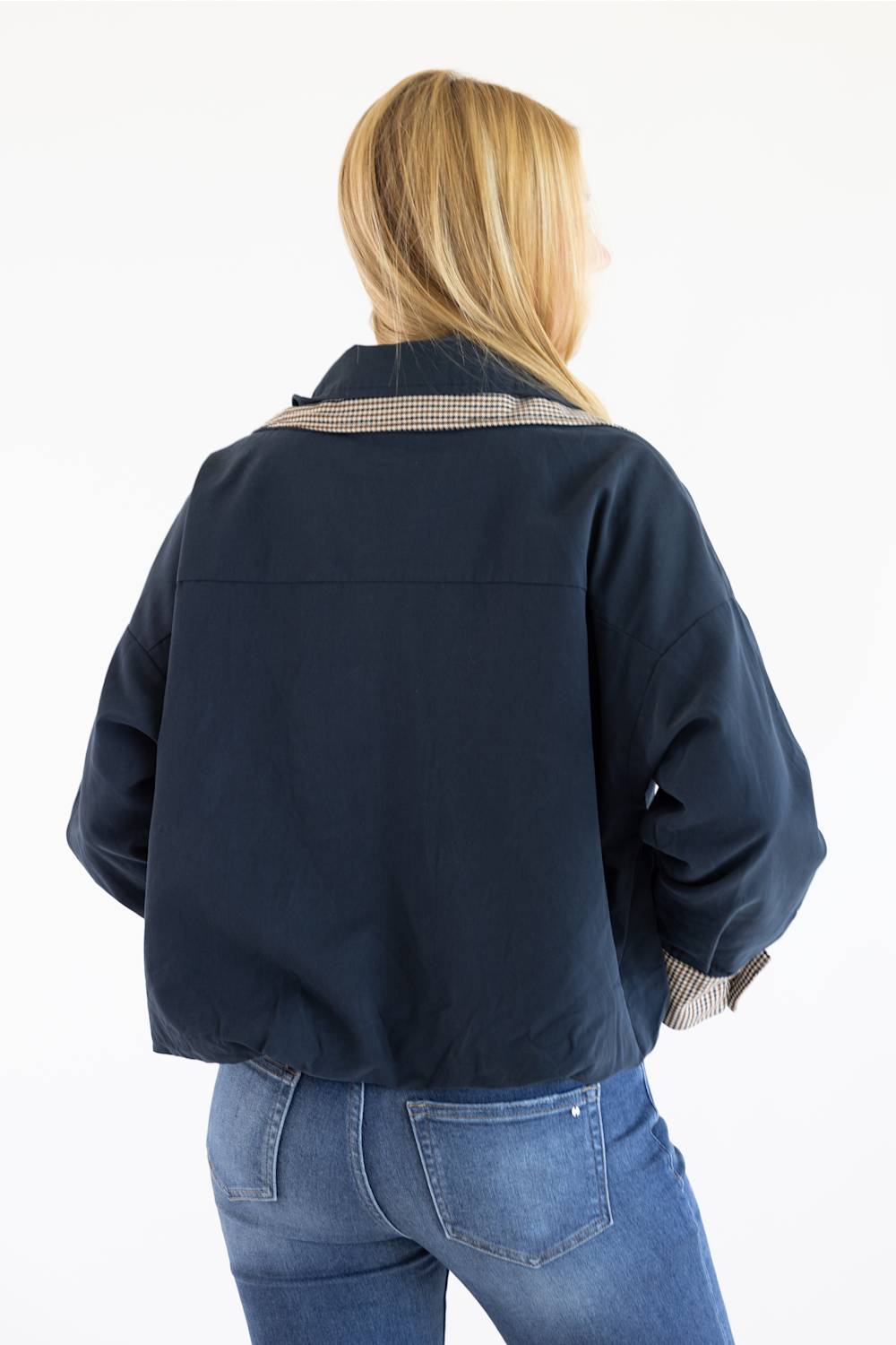 A woman viewed from behind wearing a navy blue cropped jacket with a beige-and-black checkered collar and blue jeans, against a plain white background.