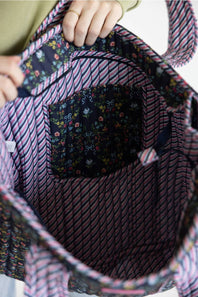 Close-up of the inside of a tote bag being held open by hands. The bag has a pink and navy striped fabric body with a floral pocket and matching striped fabric lining.