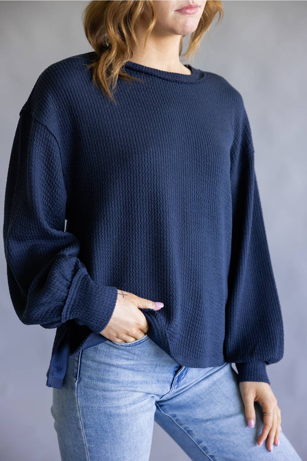 A woman wearing a navy blue waffle-knit long-sleeve sweater and light blue jeans, standing against a neutral gray studio background.