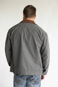 A man wearing a gray waxed cotton utility jacket with a brown corduroy collar, styled over a black t-shirt and blue jeans, standing against a light gray studio background.