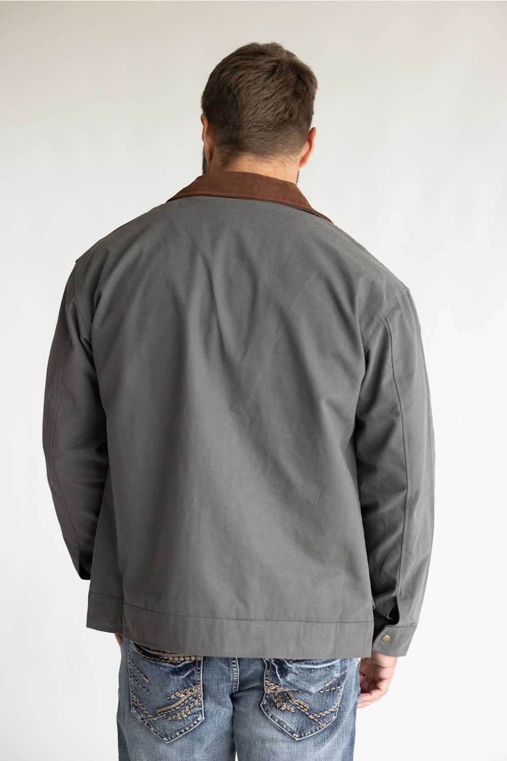 A man wearing a gray waxed cotton utility jacket with a brown corduroy collar, styled over a black t-shirt and blue jeans, standing against a light gray studio background.