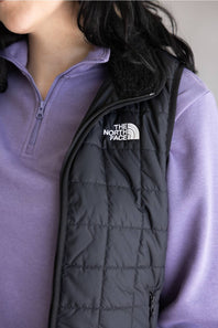 A close-up of a person wearing a black quilted puffer vest over a lavender quarter-zip pullover. The vest features a white embroidered 'The North Face' logo on the left chest and a black fleece-lined collar.