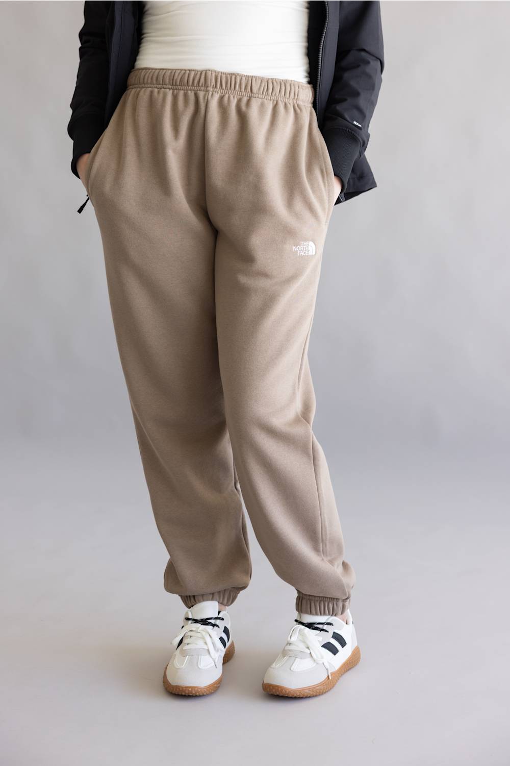 A person wearing beige jogger sweatpants with an elastic waistband and cuffs, styled with a white top, black jacket, and white sneakers with black stripes, shown from the waist down against a light gray studio background.