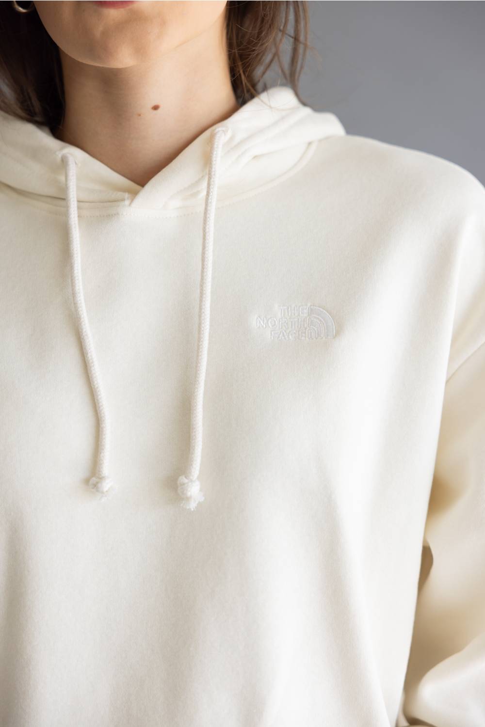 Close-up of a person wearing a cream-colored hoodie with white drawstrings and a tonal embroidered logo on the left chest, set against a neutral gray background.