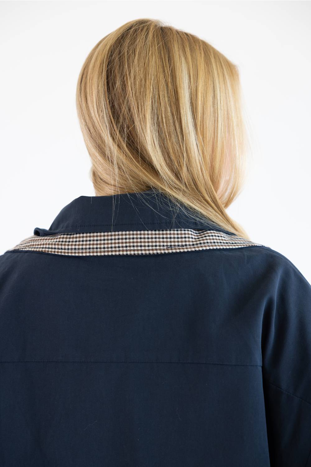 Back view of a person with blonde hair wearing a navy blue jacket with a beige and black gingham patterned collar, against a plain white background.