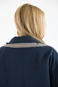 Back view of a person with blonde hair wearing a navy blue jacket with a beige and black gingham patterned collar, against a plain white background.
