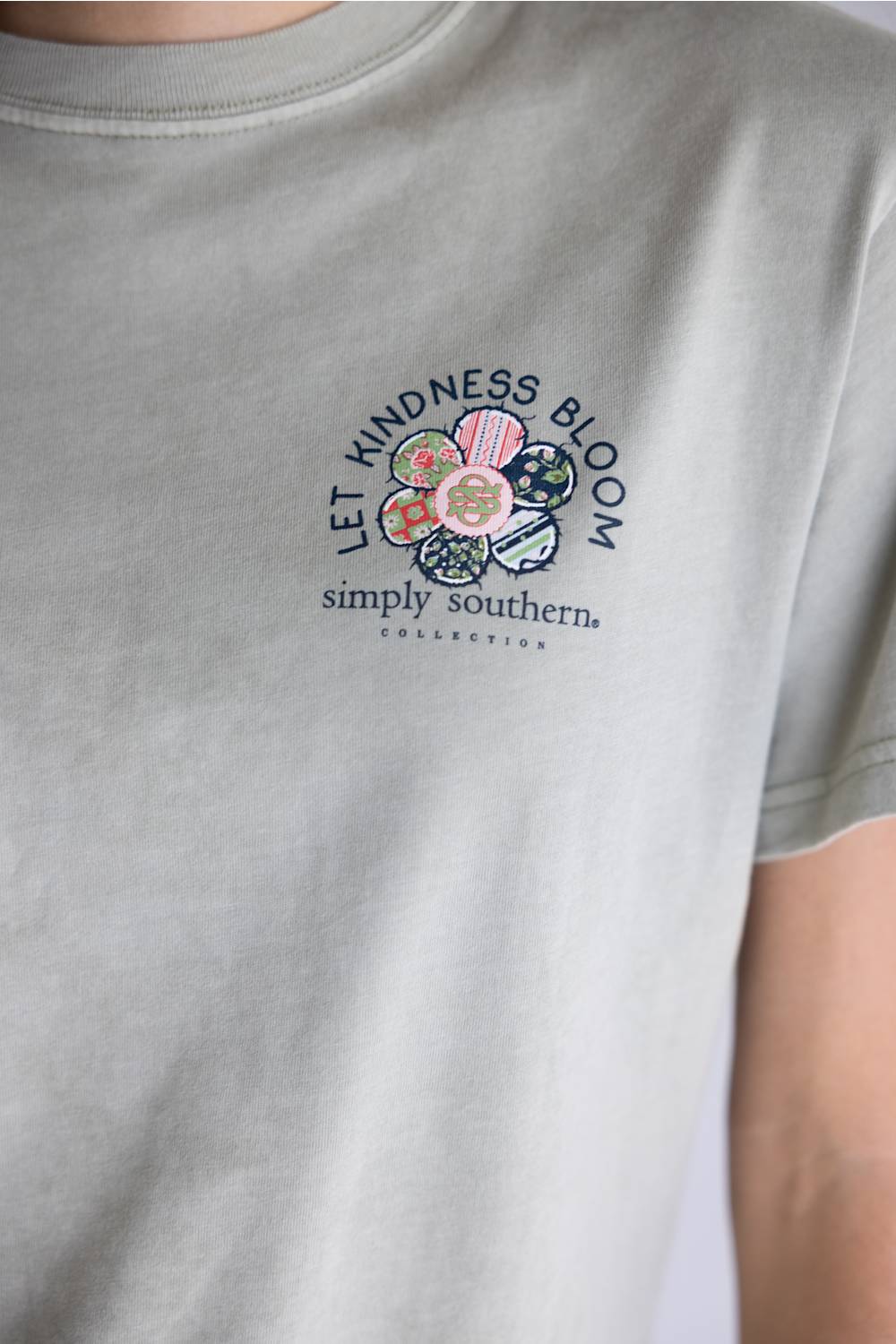 A light beige crewneck t-shirt with a colorful floral graphic on the back, featuring the text 'LET KINDNESS BLOOM' and 'simply southern.' COLLECTIONS.