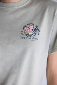 A light beige crewneck t-shirt with a colorful floral graphic on the back, featuring the text 'LET KINDNESS BLOOM' and 'simply southern.' COLLECTIONS.