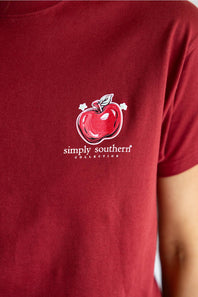 A red short-sleeve t-shirt with a large graphic print on the back, featuring a pink scalloped border, a yellow pencil, a red apple, and the text 'TEACHING IS A WORK OF ART' and 'simply southern' below it.