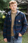 A man wearing a navy blue hooded button-up shirt jacket with red and yellow stripes, layered over a tan hoodie and blue jeans, standing outdoors in a wooded setting.