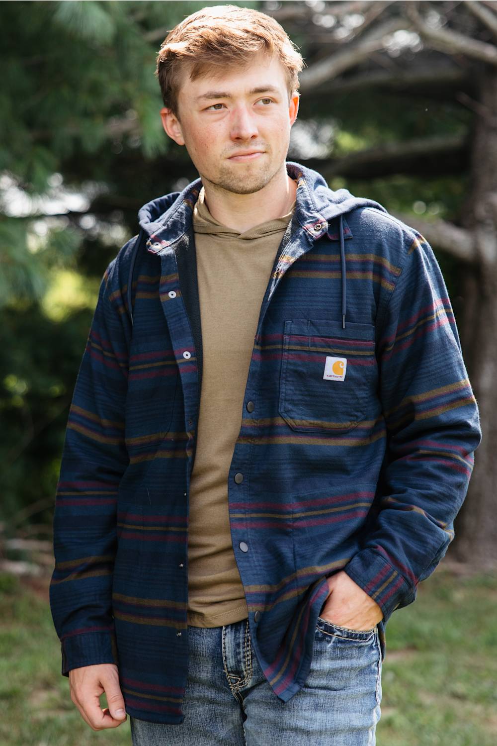 A man wearing a navy blue hooded button-up shirt jacket with red and yellow stripes, layered over a tan hoodie and blue jeans, standing outdoors in a wooded setting.