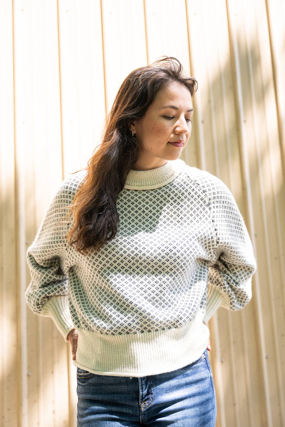 A woman wearing a cream-colored open-knit sweater with blue jeans, standing against a light wood-paneled wall.