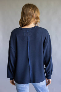 A woman viewed from the back wearing a navy blue waffle-knit long-sleeve top with a unique central seam detail and a small bow on the upper back, paired with light blue jeans against a neutral gray background.