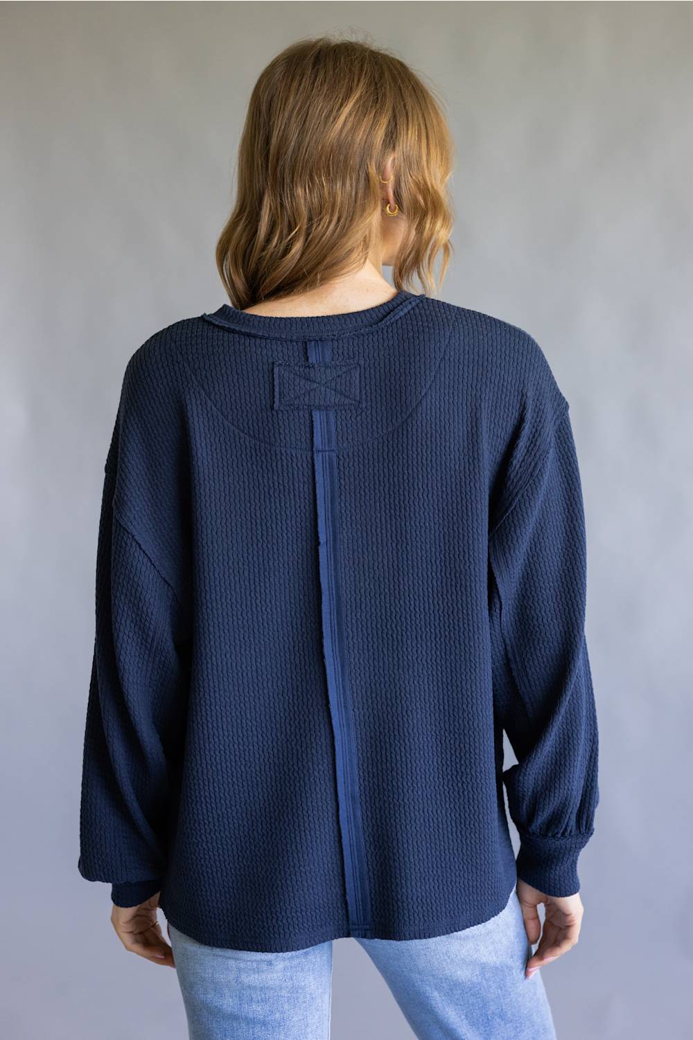 A woman viewed from the back wearing a navy blue waffle-knit long-sleeve top with a unique central seam detail and a small bow on the upper back, paired with light blue jeans against a neutral gray background.