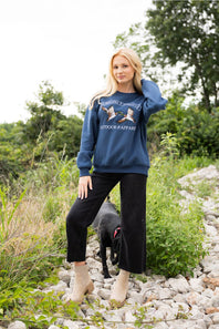 A woman wearing a navy blue crewneck sweatshirt with an embroidered graphic of two mallards and the text 'Suppliers of Quality Outdoors & Apparel'.