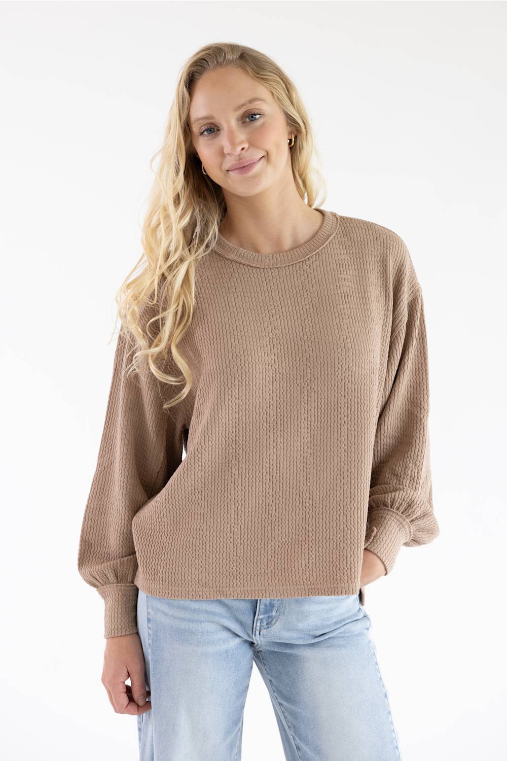 A woman with long, wavy blonde hair wearing a khaki crewneck ribbed knit top and light blue jeans against a white background.