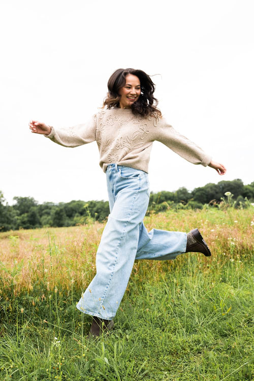 A woman running joyfully through a grassy field, wearing a beige knit sweater, light blue wide-leg jeans, and dark brown boots.